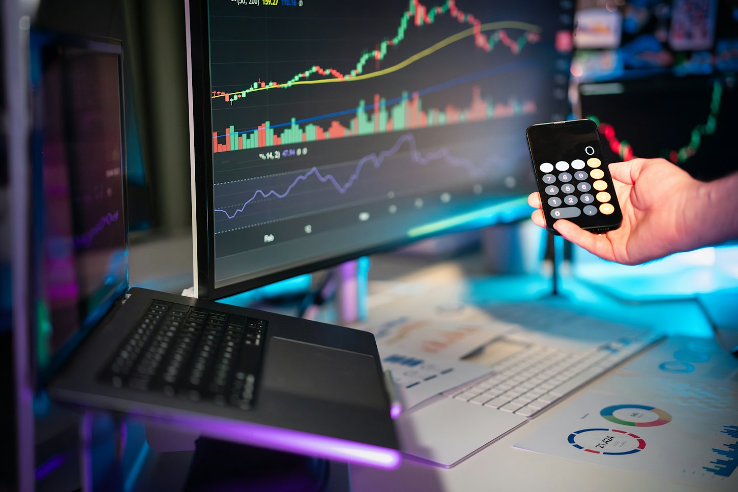 Hand hält Handy mit Taschenrechner vor Finanzdaten auf Monitoren.